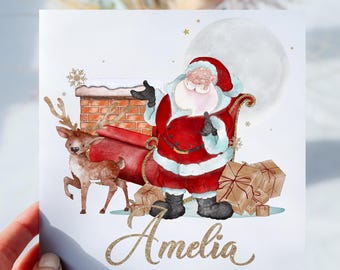 May include: A white card featuring a watercolour illustration of Santa Claus in a red suit with a white beard, delivering presents in a sleigh pulled by a reindeer. The card has a full moon in the background and the name "Amelia" written in gold glitter.