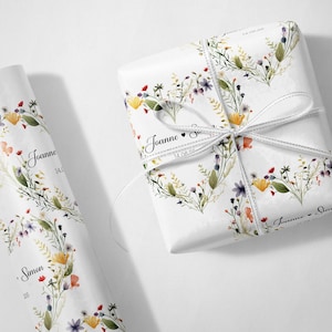 May include: White gift wrap with a floral design featuring yellow, red, and purple flowers. The wrapped gift is tied with a white ribbon. The text "Joanne" and "Simon" are printed on the wrap.