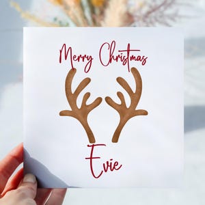May include: A white Christmas card featuring the words "Merry Christmas" in red script above brown reindeer antlers. The name "Evie" is written in red script below the antlers. The card is held by a hand.