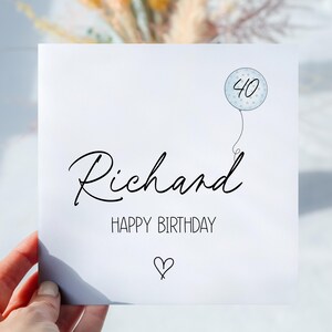 May include: A white birthday card with the name "Richard" in elegant script. Below, "HAPPY BIRTHDAY" is printed, with a small heart beneath. A balloon graphic with the number "40" floats above.