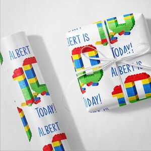 May include: White wrapping paper with a colorful block design and the text "ALBERT IS TODAY!". The design features a large number 10 constructed from red, blue, yellow, and green blocks. A white ribbon ties the wrapped gift.