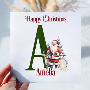 May include: A personalised Christmas card featuring a green letter A, an illustration of Santa Claus, and a reindeer. The card reads "Happy Christmas Amelia".