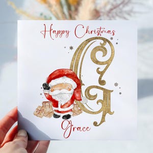 May include: A white card with gold lettering that says "Happy Christmas" and a gold letter "G" with a cartoon Santa Claus holding presents. The name "Grace" is written in red cursive at the bottom of the card.