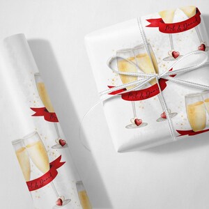 May include: White wrapping paper with a red ribbon and gold glitter. The ribbon has the text "Mil & David" on it. The paper is decorated with a repeating pattern of champagne flutes and red hearts.