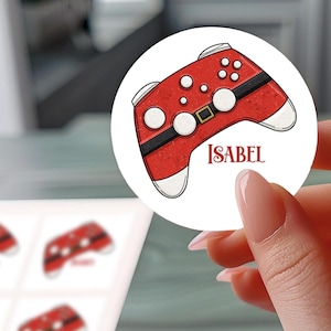 Peut inclure: Un autocollant rond avec un motif de manette rouge, des détails blancs et le nom "ISABEL". La manette a une bande noire et une boucle dorée. L'autocollant est tenu par une personne.