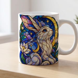 Könnte beinhalten: Ein weißer Keramikbecher mit einer farbenfrohen Illustration im Buntglas-Stil, die einen Hasen umgeben von einer Mondsichel, Sternen und einer weißen Blume zeigt. Das Design verwendet tiefe Blau-, Lila- und Goldtöne für ein lebendiges Muster.