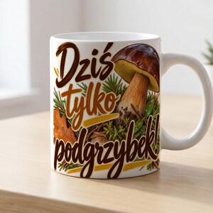 May include: A white ceramic mug featuring a brown and green graphic of a porcini mushroom surrounded by pine needles and autumn leaves. The text "Dziś tylko podgrzybek!" is printed in a dark brown script font across the front of the mug.