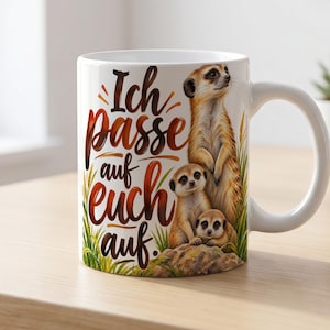 Op de afbeelding: Een witte keramische mok met een kleurrijke illustratie van drie stokstaartjes die op een rots tussen groen gras staan en zitten. De tekst "Ich passe auf euch auf." is in een bruin, gestileerd handschrift op de mok gedrukt.