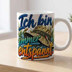 Könnte beinhalten: Eine weiße Keramiktasse mit einer Grafik eines beigen Anglerhutes mit einem Angelköder, umgeben von grünen Schilfhalmen und Wasserspritzern. Der Text "Ich bin immer entspannt" ist in blauer, grüner und orangefarbener Schrift auf der Vorderseite.