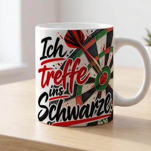 May include: A white ceramic mug featuring a graphic of a dartboard with a red and black dart hitting the center. The text Ich treffe ins Schwarze is printed in a mix of black and red script and bold lettering across the front of the mug.