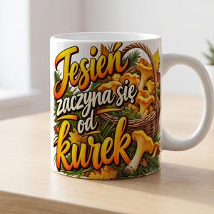 Könnte beinhalten: Ein weißer Keramikbecher mit einer Grafik eines geflochtenen Korbs voller gelber Pfifferlinge, grüner Tannennadeln und Herbstblätter. Der Text "Jesień zaczyna się od kurek" ist in großen, stilisierten orangefarbenen und schwarzen Buchstaben gedruckt.