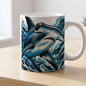 Pode incluir: Uma caneca de cerâmica branca com uma ilustração gráfica detalhada de um tubarão-martelo a nadar em água azul com peixes pequenos e bolhas. A obra utiliza tons de azul escuro e claro com linhas pretas para criar profundidade.