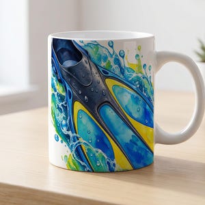 Puede incluir: Una taza de cerámica blanca con una vibrante ilustración en acuarela de una aleta de natación negra, azul y amarilla rodeada de salpicaduras de agua. El diseño envuelve el exterior, mostrando un tema acuático dinámico con colores vivos.