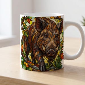 Könnte beinhalten: Ein weißer Keramikbecher mit einer Illustration im Buntglas-Stil, die ein braunes Wildschwein mit markanten Stoßzähnen zeigt. Das Design umfasst herbstlich gefärbte Blätter, Eicheln und grünes Laub vor einem gelben, geometrischen Hintergrund.