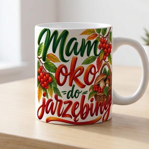 Puede incluir: Una taza de cerámica blanca con una ilustración colorida de bayas de serbal rojas, hojas verdes y una cesta tejida llena de setas marrones. El texto "Mam oko do jarzębiny" está impreso en una fuente cursiva estilizada, negrita y multicolor.