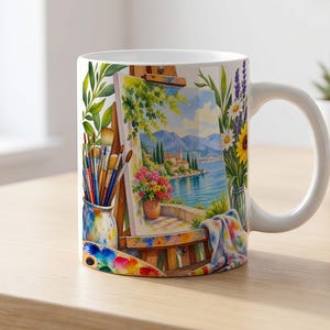May include: A white ceramic mug featuring a colorful illustration of an artist's easel holding a painting of a coastal landscape with mountains, a lake, and flowers. The design includes paintbrushes in a jar, a palette, and floral arrangements.