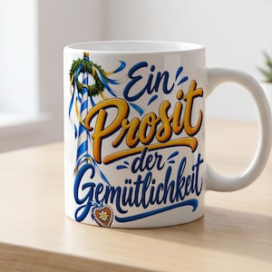 Pode incluir: Uma caneca de cerâmica branca com um gráfico azul e amarelo de um mastro de maio com uma coroa e o texto Ein Prosit der Gemütlichkeit em letras azuis e amarelas. Na base, há um pequeno biscoito de gengibre em forma de coração.