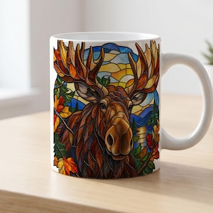 May include: A white ceramic mug featuring a stained glass style illustration of a brown moose with large antlers. The background depicts a landscape with autumn leaves, green trees, blue mountains, and a yellow sun, creating a colorful scene.