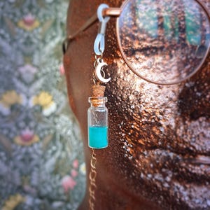 May include: A gold chain with a small glass bottle filled with turquoise liquid and a crescent moon charm. The chain is attached to a pair of glasses.