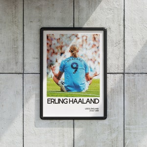 May include: A black and white poster with a photo of a soccer player sitting in a lotus position on a green field. The player is wearing a light blue jersey with the number 9 and the name "HAALAND" on the back. The text "ERLING HAALAND" is printed in black at the bottom of the poster, along with the text "LEEDS, ENGLAND 21 JULY 2000".