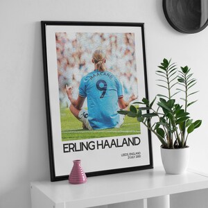 May include: A black framed poster with a photo of a soccer player in a blue jersey with the number 9 on the back. The player is sitting in a meditative pose on a green field. The text "ERLING HAALAND" is printed in white below the photo. The text "LEEDS, ENGLAND 21 JULY 2000" is printed in white below the text "ERLING HAALAND".