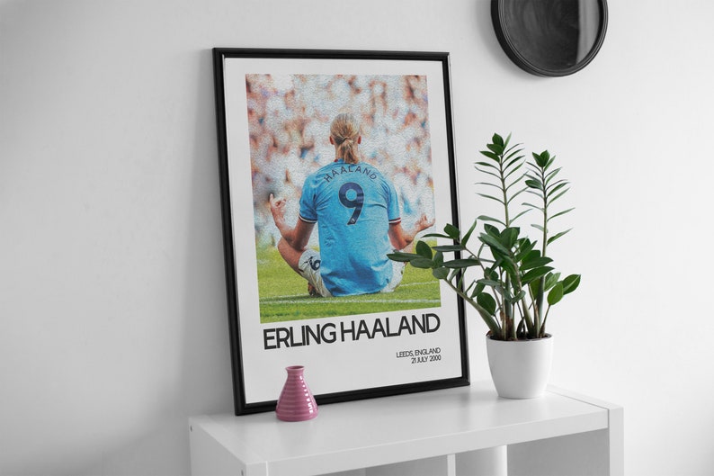 May include: A black framed poster with a photo of a soccer player in a blue jersey with the number 9 on the back. The player is sitting in a meditative pose on a green field. The text "ERLING HAALAND" is printed in white below the photo. The text "LEEDS, ENGLAND 21 JULY 2000" is printed in white below the text "ERLING HAALAND".
