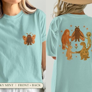 May include: A light mint green t-shirt featuring a small moth graphic on the front left chest. The back displays a large illustration of a moth, a sasquatch, an alien, a jackalope, and a dinosaur holding hands in a circle with small stars.