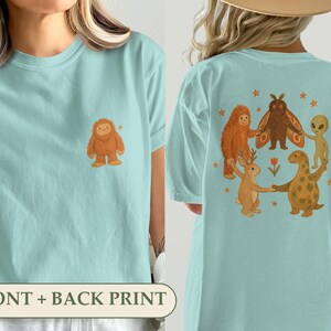May include: A light blue t-shirt featuring a small brown creature graphic on the front left chest. The back displays a large circular illustration of a brown creature, a moth, an alien, a jackalope, and a dinosaur holding hands with stars.