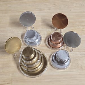 May include: A collection of circular metal lids in various sizes and finishes, including gold, silver, copper, and brushed steel. Some lids are stacked in nested sets, while others are displayed upright on small clear plastic stands.