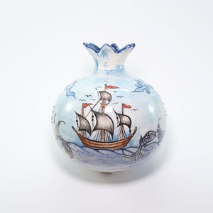 May include: A ceramic vase shaped like a pomegranate, painted with a light blue background and a detailed illustration of a sailing ship. The vase has a decorative, scalloped rim and floral accents.