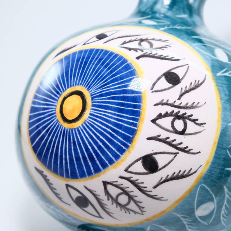 May include: A teal ceramic vase with a white and yellow circular design featuring a blue sunburst pattern and multiple black and white eye illustrations. The vase has a rounded shape and a narrow neck.