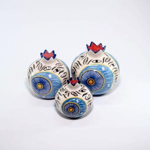 May include: Three ceramic pomegranate sculptures. The pomegranates are white with blue and yellow circular designs, and black eye patterns. Each has a red and blue crown. The sculptures are of varying sizes.