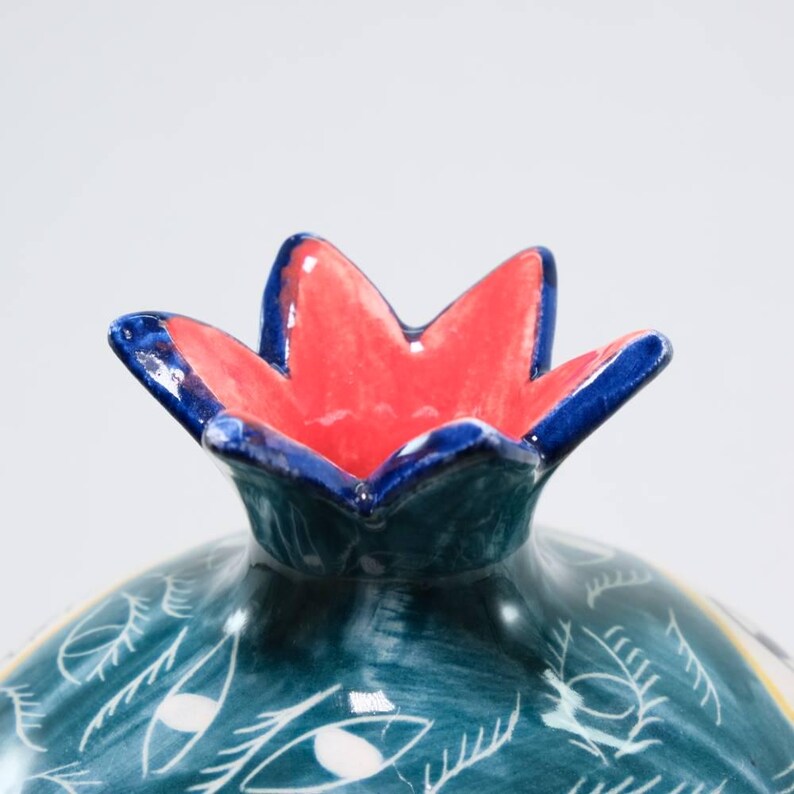May include: A ceramic pomegranate-shaped container with a red and blue flower-like top. The body is teal with white eye and leaf patterns. The container is likely used for decorative purposes.