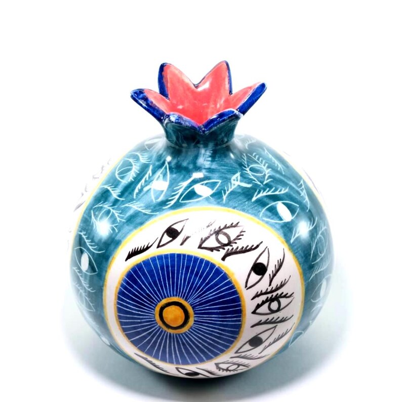 May include: A ceramic pomegranate with a teal base, adorned with white eye and leaf patterns. The design includes a central blue and yellow circular motif, and a red and blue crown. This decorative piece is likely a unique home decor item.