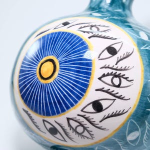May include: A teal ceramic vase with a white and yellow circular design featuring a blue sunburst pattern and black eye illustrations. The vase has a rounded shape and a narrow neck, with white leaf-like patterns.