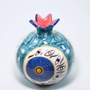 May include: A ceramic pomegranate with a teal base, adorned with white eye and leaf patterns. The design includes a central blue and yellow circular motif. The top is red with blue accents.
