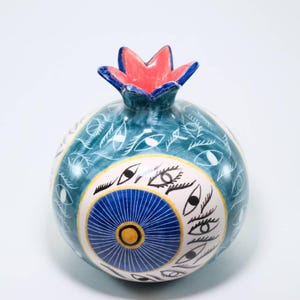 May include: A ceramic pomegranate sculpture with a teal base, adorned with white eye motifs and a blue and yellow circular design. The top features a red and blue crown. This decorative piece is ideal for home decor.