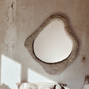 May include: A bedroom scene featuring a bed with a light-colored comforter and pillows. A unique, irregularly shaped mirror hangs on the wall. A wooden trunk serves as a nightstand, holding books and a glass of water. A potted plant adds a touch of greenery.