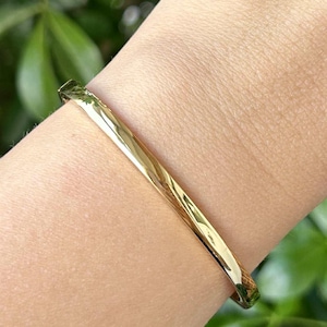 May include: A gold bangle bracelet. The bracelet is a solid band with a smooth, reflective surface. The jewellery is worn on a wrist, with a blurred green background.