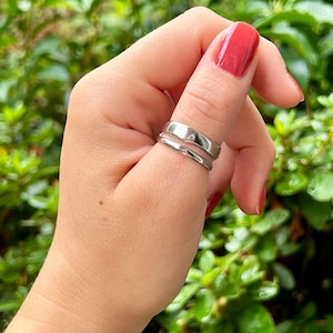 May include: A close-up of a hand wearing two silver rings on the thumb. The hand is against a backdrop of green foliage.