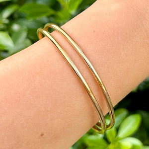 May include: Two gold-colored bangles are worn on a person's arm. The bangles are smooth and shiny, reflecting light. The background is blurred green foliage.