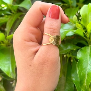 May include: A gold thumb ring with a hammered texture and a circular design. The ring is worn on a person's thumb.