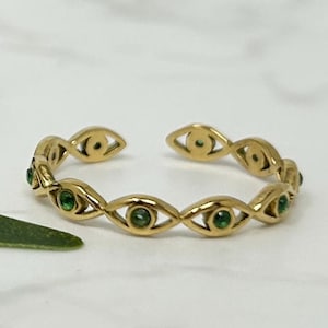 May include: A gold ring with an open design, featuring a series of eye-shaped links. Each eye shape is accented with a small, round, green gemstone. The ring is set against a white background.