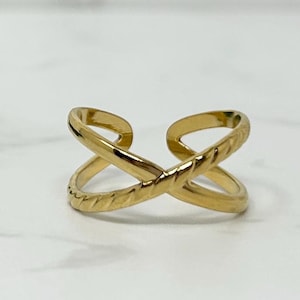 May include: A gold-colored, open-ended ring with an X-shaped design. The ring features a textured, rope-like pattern on the crossing bands. The ring is displayed on a white surface.