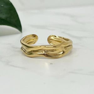 Puede incluir: Un anillo de color dorado con un diseño ondulado y texturado. El anillo es abierto y ajustable.
