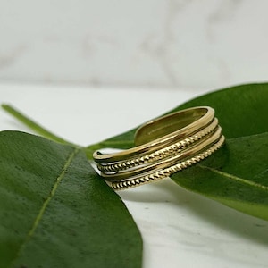 May include: A gold-colored ring with a layered design. The ring features three bands with textured detailing, including a beaded pattern. The ring is open at the back and rests on a green leaf.
