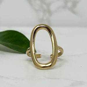 May include: A gold-coloured ring featuring an open oval design. The ring has a smooth, polished finish and a minimalist style. The ring is set against a white marble surface with a green leaf in the background.