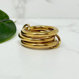 May include: A gold-colored ring with a multi-loop design. The ring features four connected, rounded bands, creating a stacked appearance. The ring is made of a shiny, reflective metal and sits on a white surface. A green leaf is visible in the background.