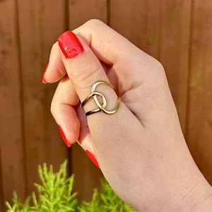May include: A gold-colored ring with an intertwined design. The ring is worn on a finger with red nail polish. The background features a wooden fence and green foliage.