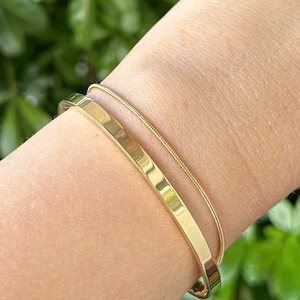May include: Two gold bracelets on a wrist. One is a thick, polished bangle, and the other is a delicate chain. The bracelets are set against a background of green foliage. The jewelry is simple and elegant.